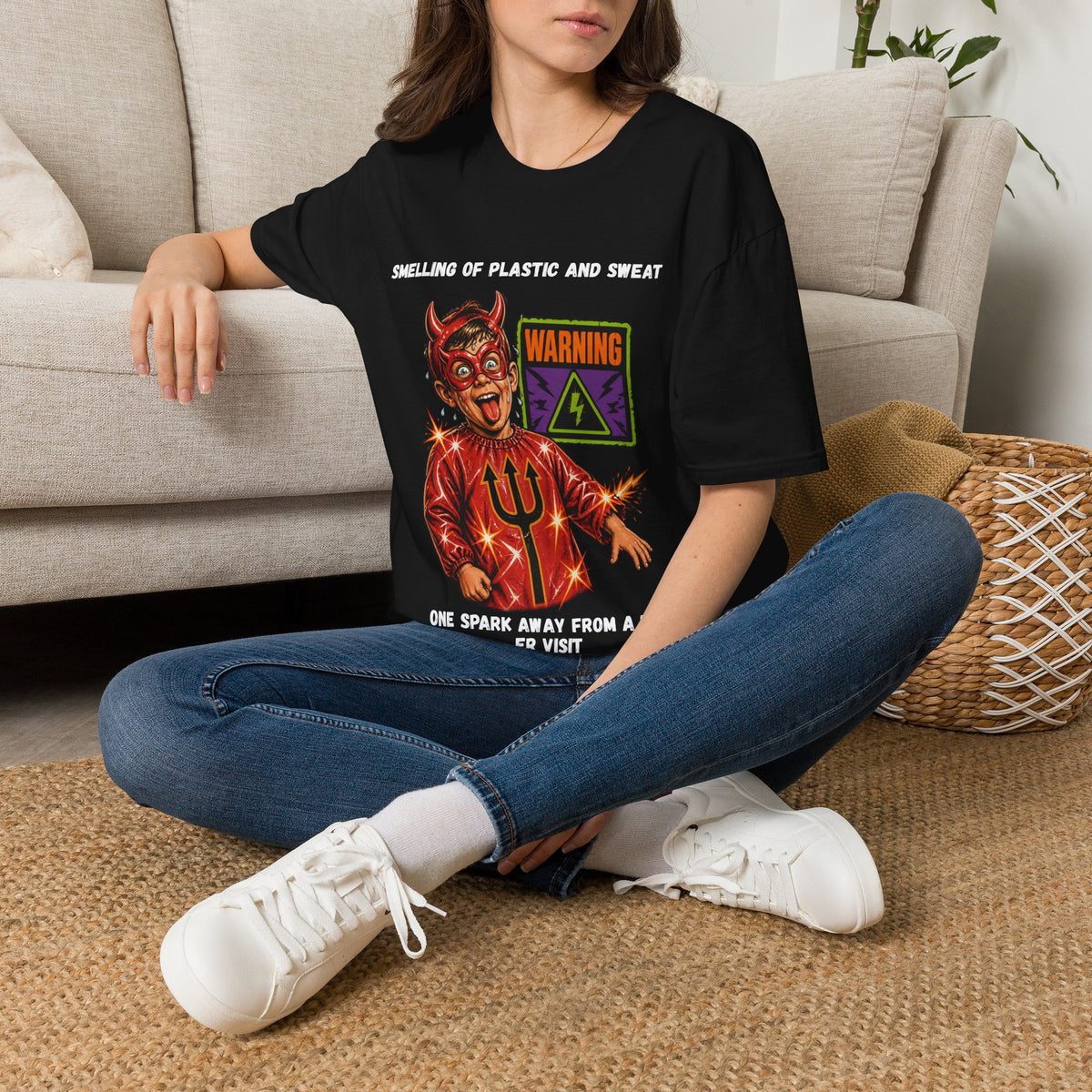 A woman sitting cross-legged on a woven rug in front of a beige sofa, wearing a black graphic T-shirt with a red devil illustration and text, blue jeans, and white sneakers.
