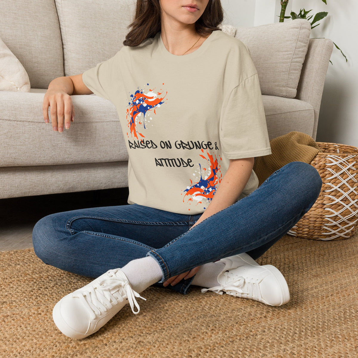 A woman sitting cross-legged on a woven rug in front of a beige sofa, wearing a sand graphic T-shirt with blue jeans and white sneakers. The T-shirt features a colorful paint-splatter design and the text 'RAISED ON GRUNGE & ATTITUDE'.