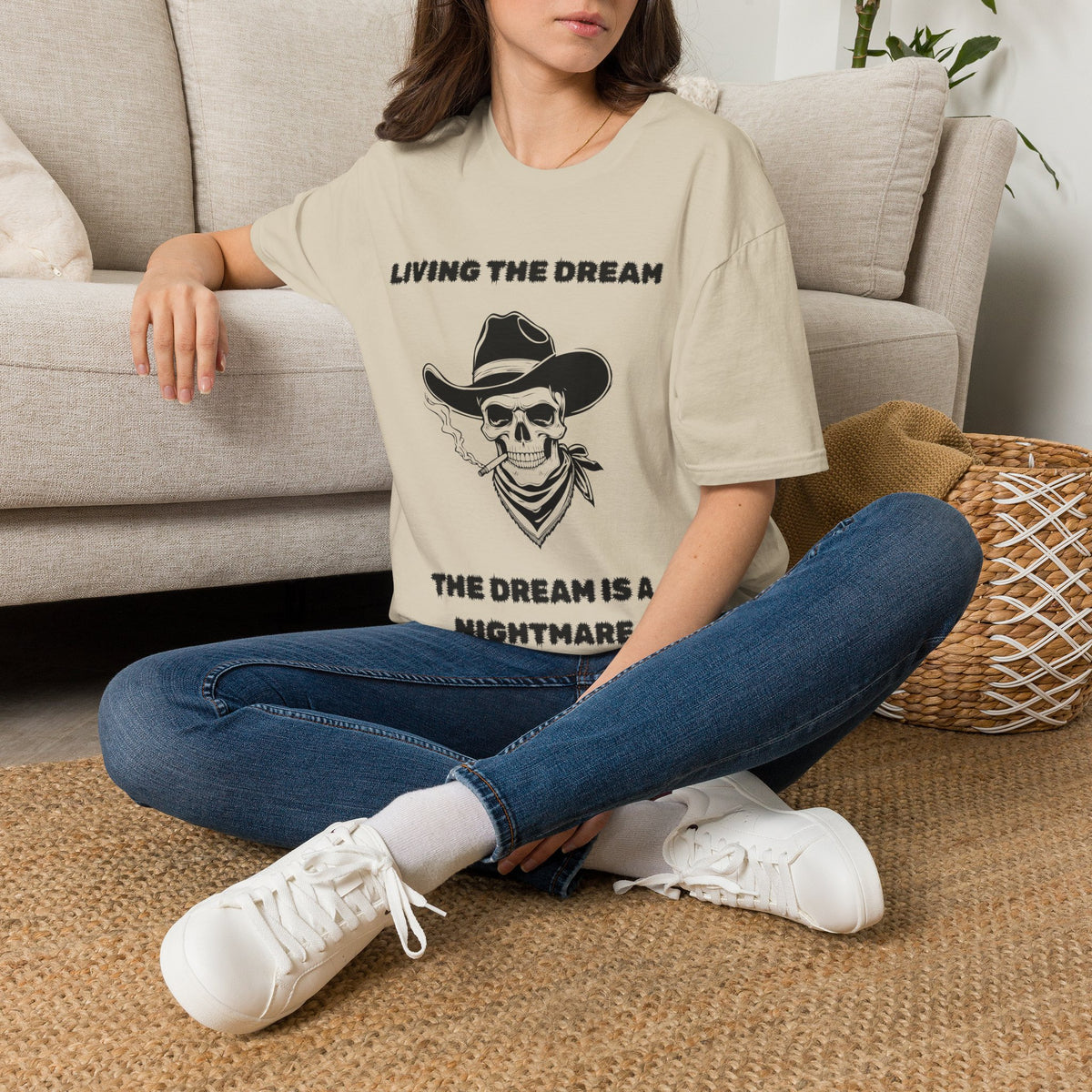 A woman sitting cross-legged on a woven rug in front of a light beige sofa, wearing a sand graphic t-shirt with a black skull and cowboy hat design, blue jeans, and white sneakers.