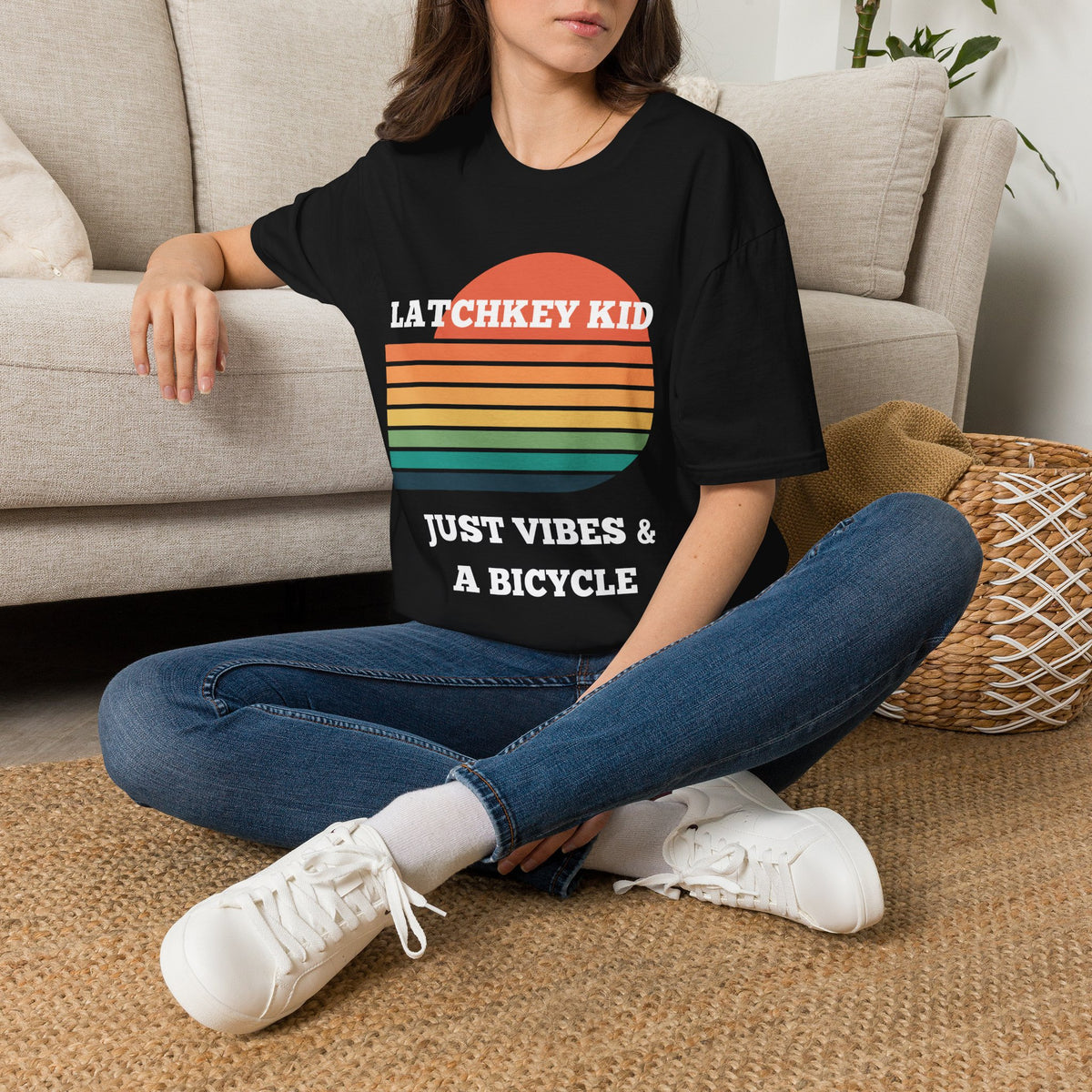 A woman sitting on a woven rug in front of a beige couch, wearing a black graphic t-shirt with a retro sunset design and the text 'LATCHKEY KID JUST VIBES & A BICYCLE'.