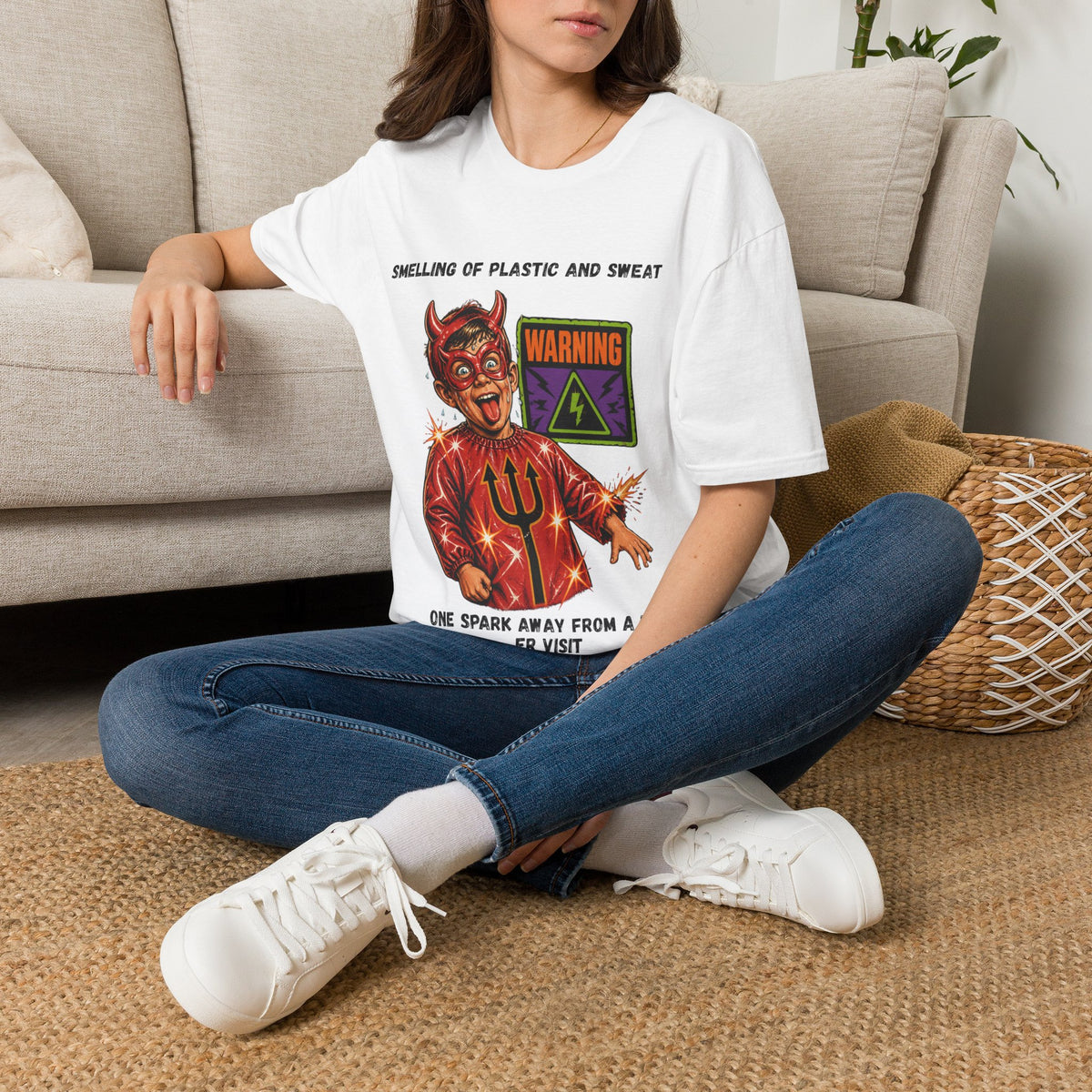 A woman sitting cross-legged on a woven rug in front of a beige sofa, wearing a white graphic t-shirt with a devil design and text, blue jeans, and white sneakers.