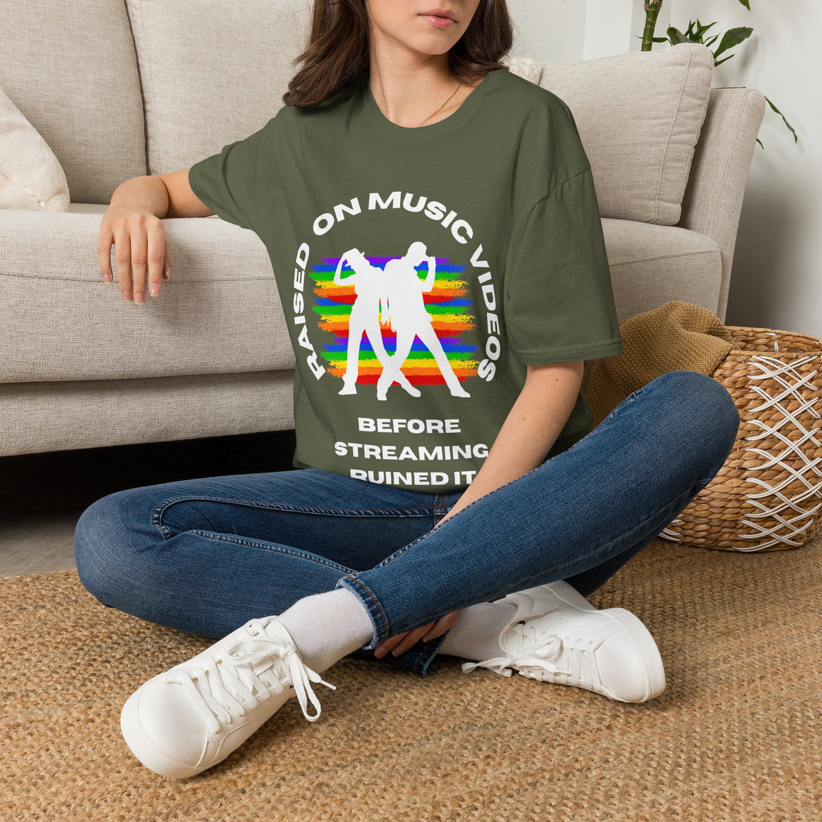 A woman sitting cross-legged on a woven rug in front of a beige sofa, wearing an green graphic T-shirt with white text and a rainbow-striped silhouette design, paired with blue jeans and white sneakers.