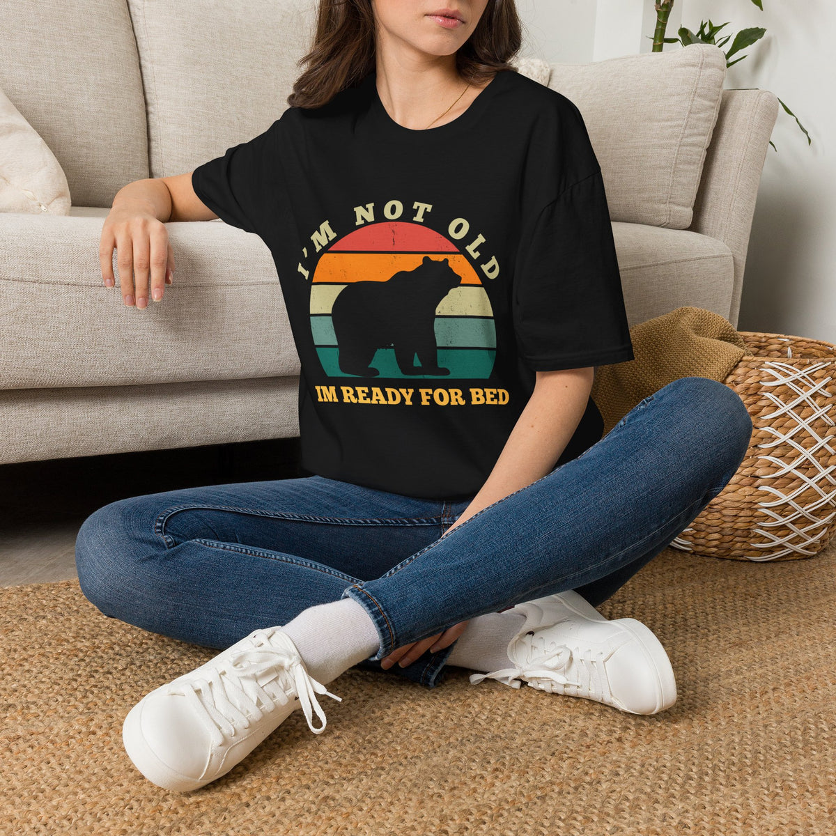 A woman sitting cross-legged on a woven rug in front of a beige sofa, wearing a black graphic T-shirt with a retro sunset bear design and the text 'I'M NOT OLD I'M JUST TIRED', paired with blue jeans and white sneakers.