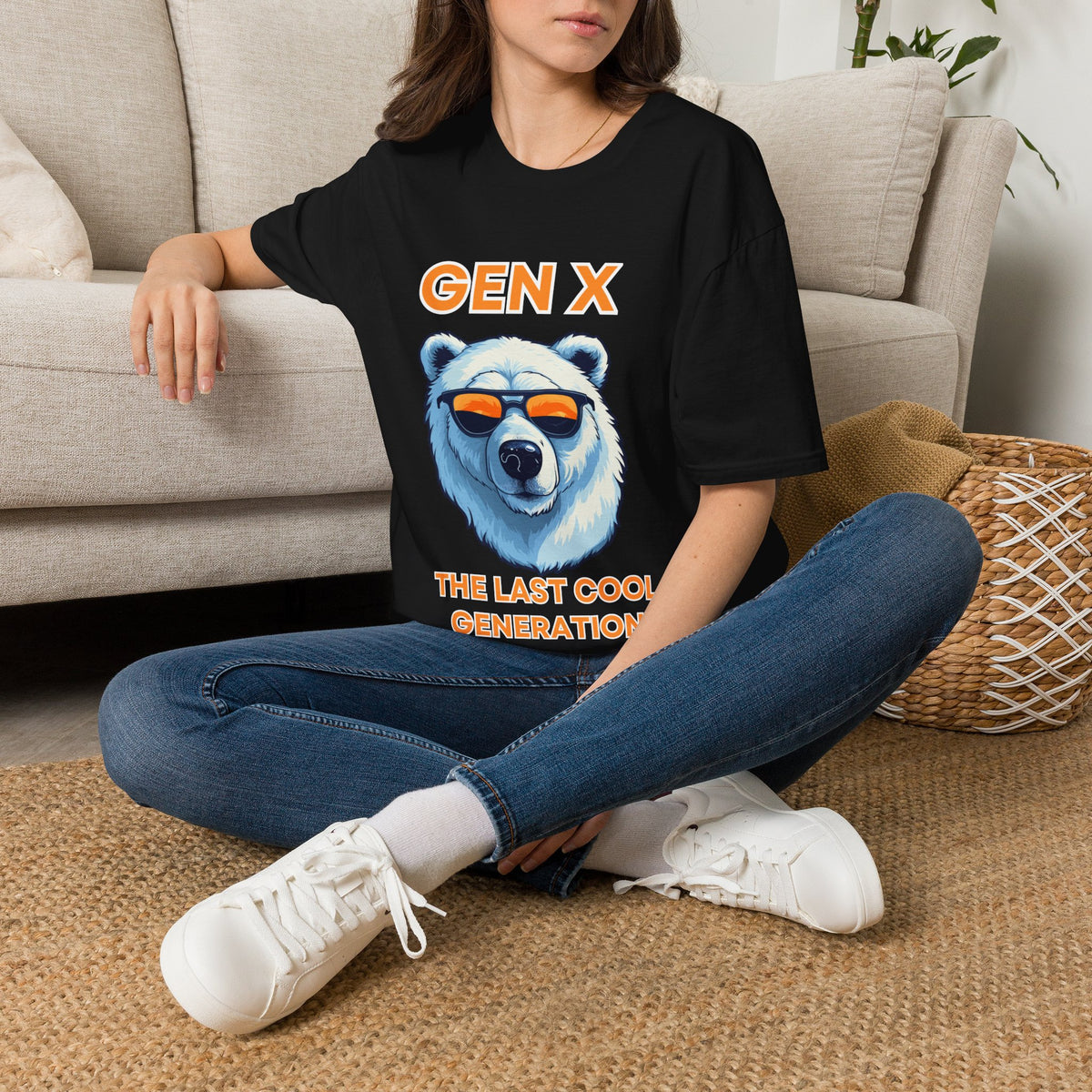 A woman sitting cross-legged on a woven rug in front of a beige sofa, wearing an oversized black graphic T-shirt with a polar bear wearing orange sunglasses and the text 'GEN X THE LAST COOL GENERATION'.