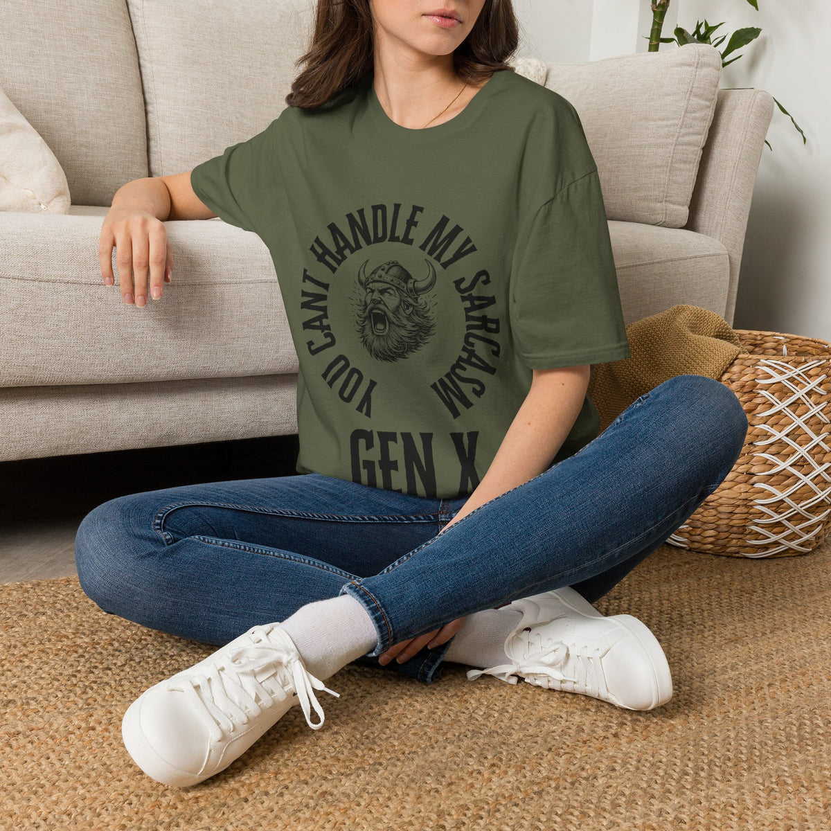 A woman sat on rug next to a couch wearing a green short-sleeve graphic t-shirt with a black circular print featuring a roaring viking man and the text 'YOU CAN'T HANDLE MY SARCASM GEN X'. 