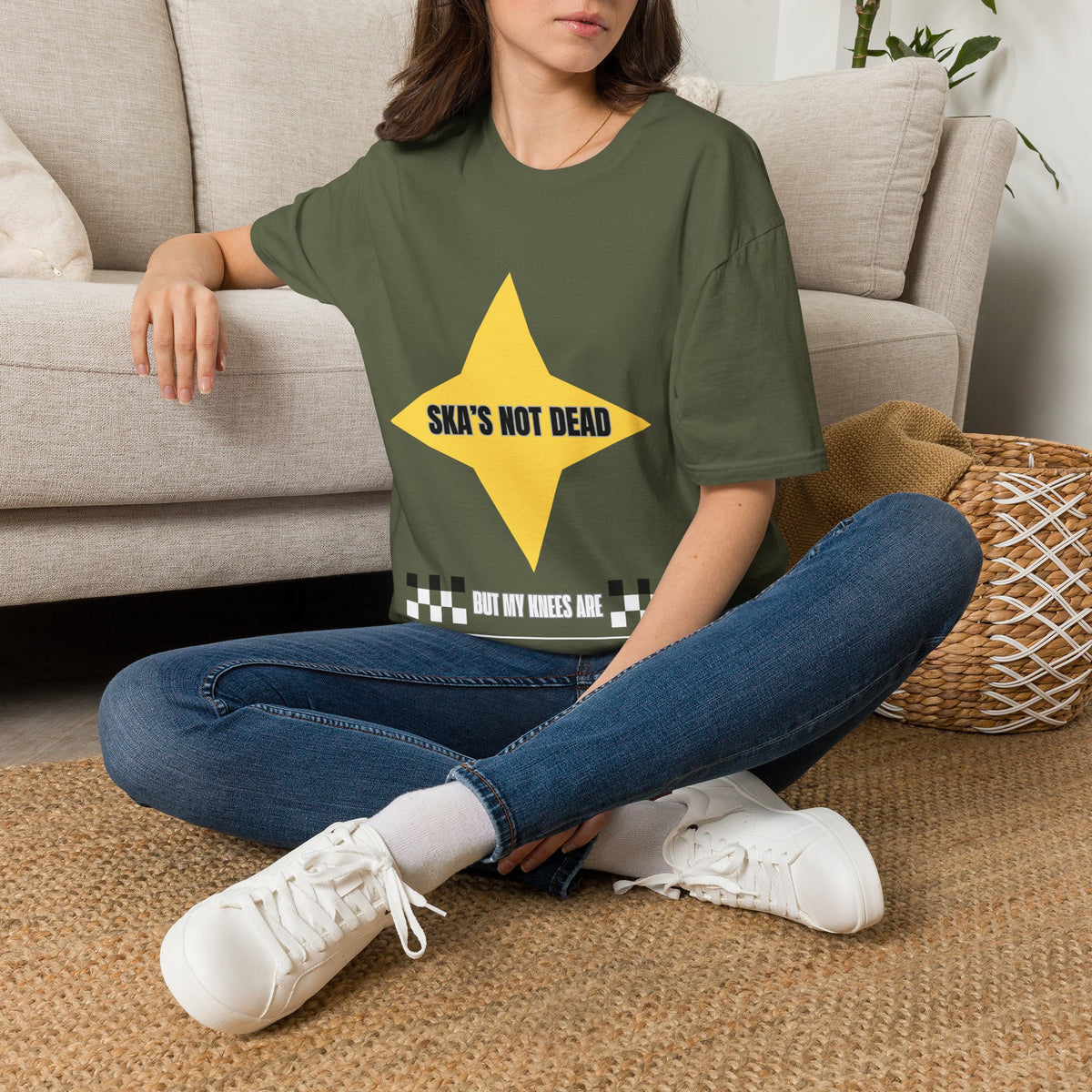 A woman sitting on a woven rug in front of a beige sofa, wearing a green graphic t-shirt with a yellow star and the text 'SKA'S NOT DEAD BUT MY KNEES ARE'.