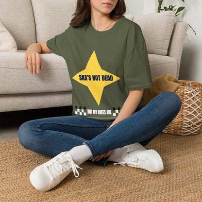 A woman sitting on a woven rug in front of a beige sofa, wearing a green graphic t-shirt with a yellow star and the text 'SKA'S NOT DEAD BUT MY KNEES ARE'.