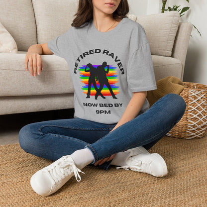 A woman sitting cross-legged on a woven rug in front of a beige sofa, wearing a gray graphic T-shirt with a rainbow design and the text 'RETIRED RAVEN NOW BED BY 9PM'.
