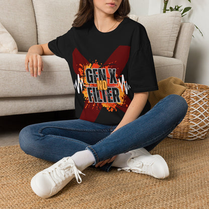 A woman sitting cross-legged on a woven rug in front of a beige sofa, wearing a black graphic T-shirt with a large red 'X'and text of 'GEN X NO FILTER' design, blue jeans, and white sneakers.