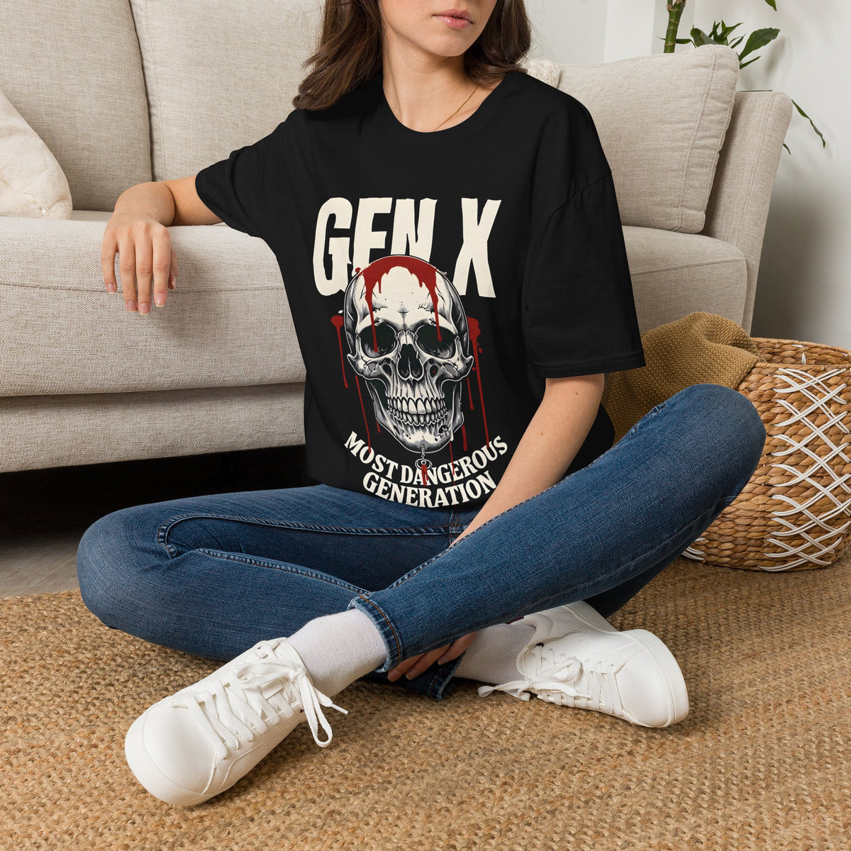 A woman sitting cross-legged on a woven rug in front of a beige sofa, wearing a black graphic T-shirt with the text 'GEN X MOST DANGEROUS GENERATION' and a skull with dripping blood illustration, paired with blue jeans and white sneakers.