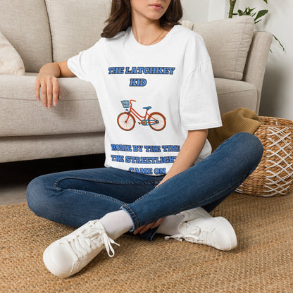 A woman sitting on a woven rug in front of a beige couch, wearing a white graphic t-shirt with blue text reading 'THE LATCHKEY KID' and 'HOME BY THE TIME THE STREETLIGHT GAVE ON' around an illustration of a red bicycle.