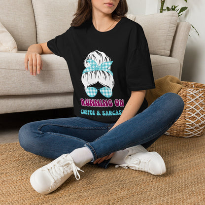 A woman sitting on a woven rug in front of a beige sofa, wearing a black graphic t-shirt with a cartoon hair bun and bow design above the text 'RUNNING ON COFFEE & SARCASM'.