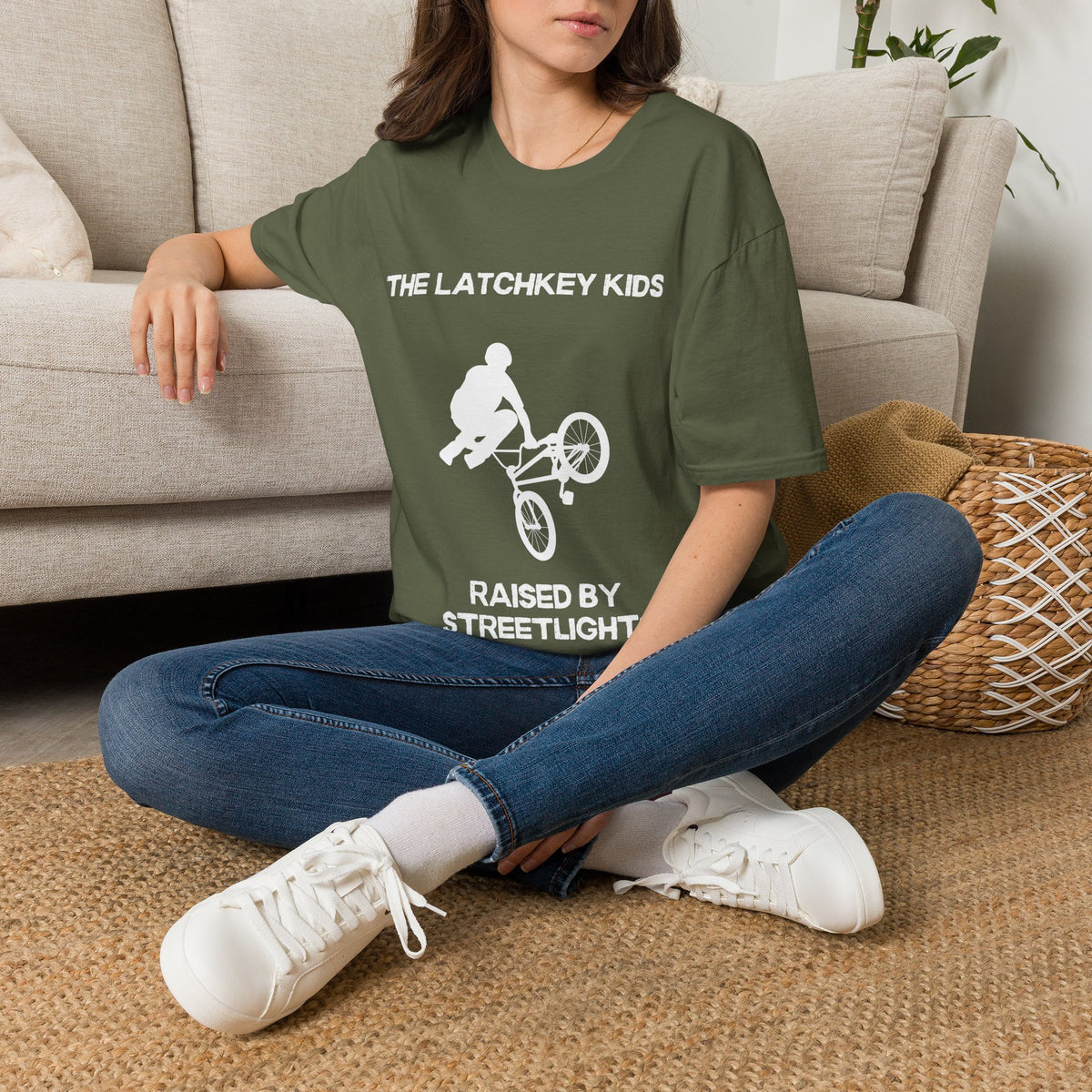A woman sitting cross-legged on a woven rug in front of a beige sofa, wearing a green graphic T-shirt with white text reading 'THE LATCHKEY KIDS' and 'RAISED BY STREELIGHT' around a BMX rider illustration, paired with blue jeans and white sneakers.