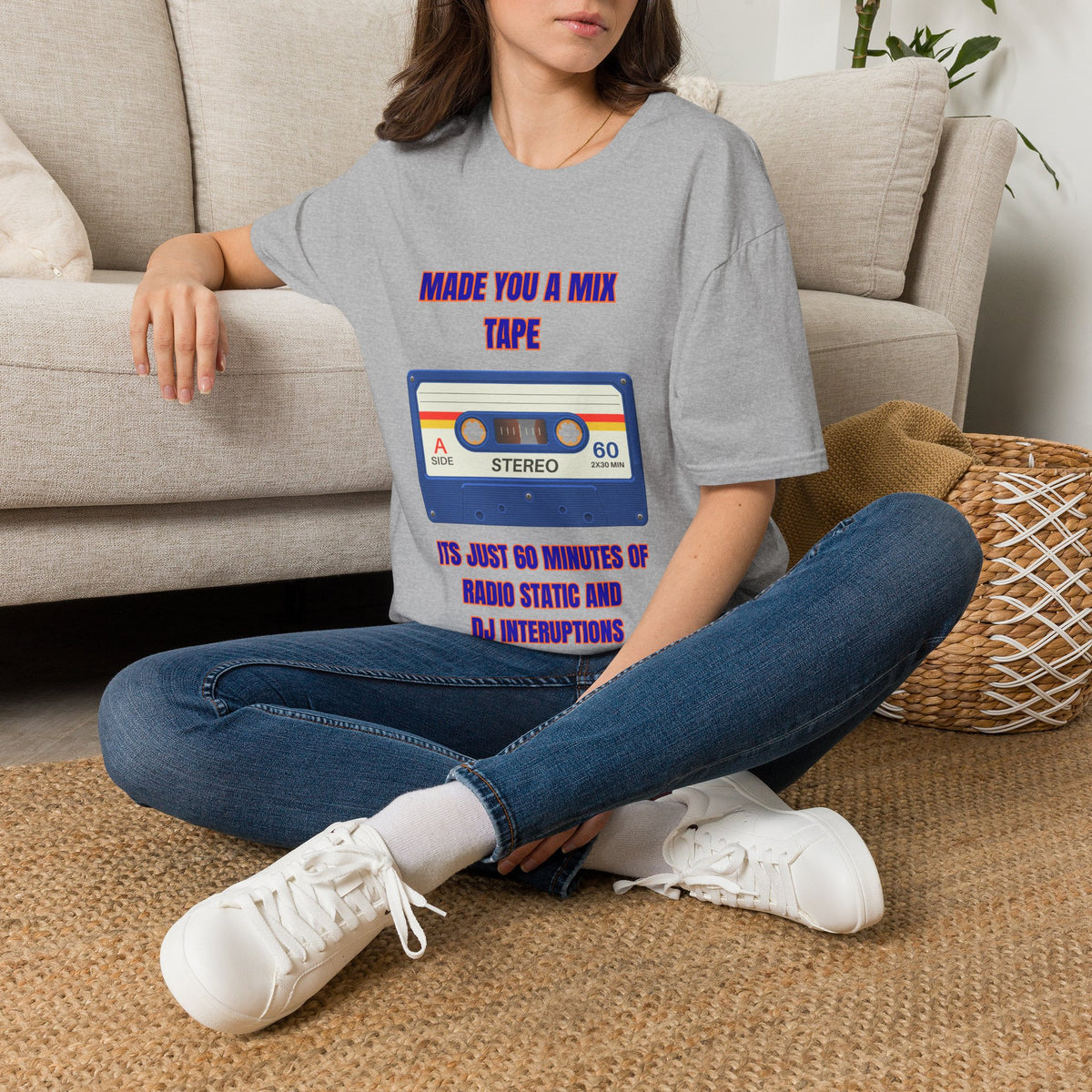 A woman sits cross-legged on a woven rug in front of a beige sofa, wearing a gray graphic T-shirt with a retro cassette tape design and purple text that reads 'MADE YOU A MIX TAPE' and 'ITS JUST 60 MINUTES OF RADIO STATIC AND DJ INTERRUPTIONS.'.