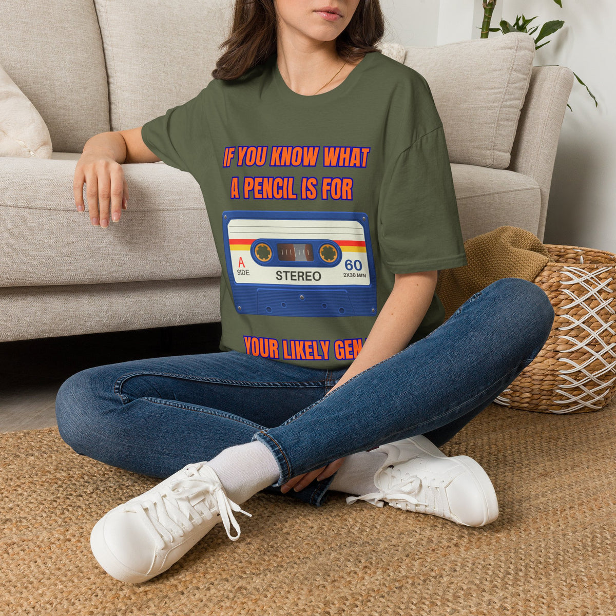 A woman sitting on a woven rug in front of a beige couch, wearing a green graphic t-shirt with a retro cassette tape design and red text that reads 'IF YOU KNOW WHAT A PENCIL IS FOR YOUR LIKELY GENIUS'.