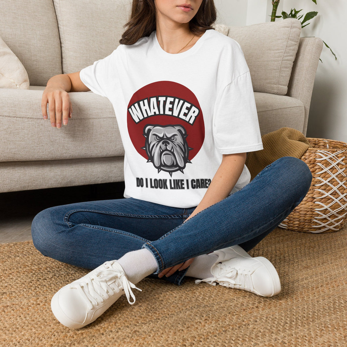 A woman sitting ona  rug next to a couch wearing a white t-shirt. The shirt has a red circle with a bulldog on it and the text that reads " WHATEVER, DO I LOOK LIKE I CARE"