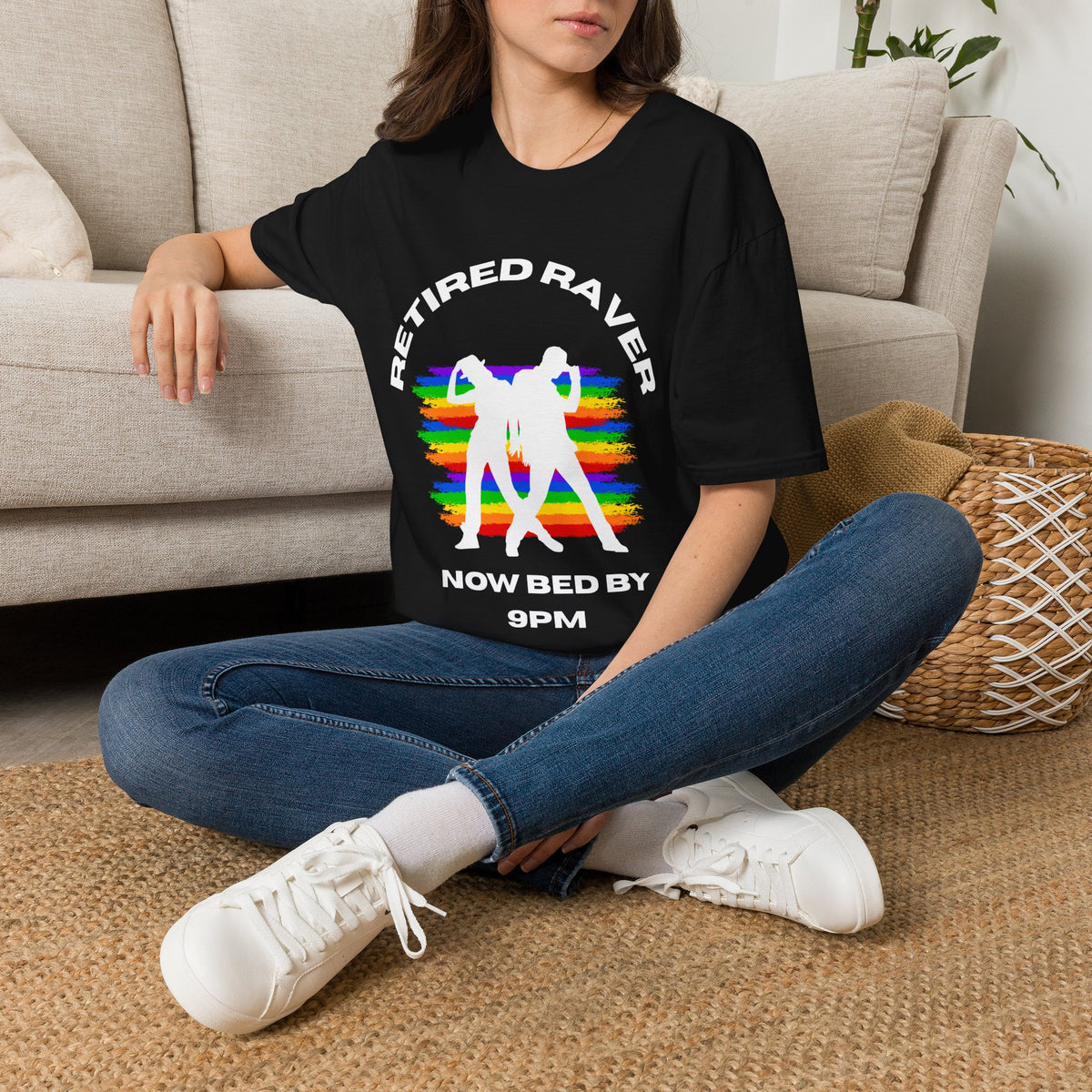 Person sitting on a carpeted floor wearing a black graphic t-shirt with text "REITIRED RAVERS NOW BED BY 9PM" with rainbow colours and white dancer silhouette design.