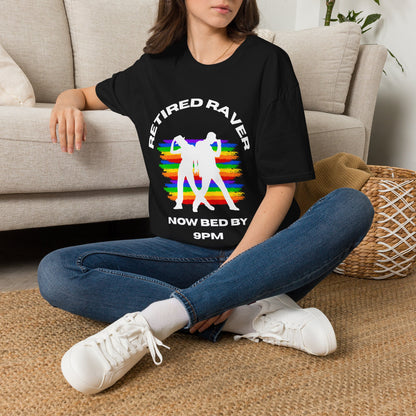 Person sitting on a carpeted floor wearing a black graphic t-shirt with text "REITIRED RAVERS NOW BED BY 9PM" with rainbow colours and white dancer silhouette design.