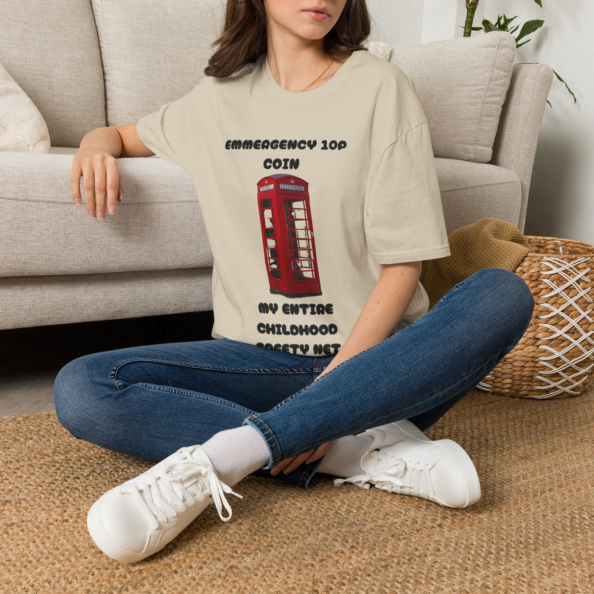 A woman sat on a rug next to a couch wearing a sand graphic t-shirt displayed on a rustic wood background, featuring a red telephone booth graphic and the text 'EMERGENCY 10P COIN MY ENTIRE CHILDHOOD SAFETY NET'. 