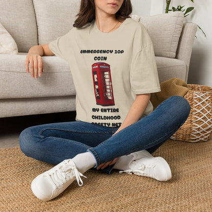 A woman sat on a rug next to a couch wearing a sand graphic t-shirt displayed on a rustic wood background, featuring a red telephone booth graphic and the text 'EMERGENCY 10P COIN MY ENTIRE CHILDHOOD SAFETY NET'. 