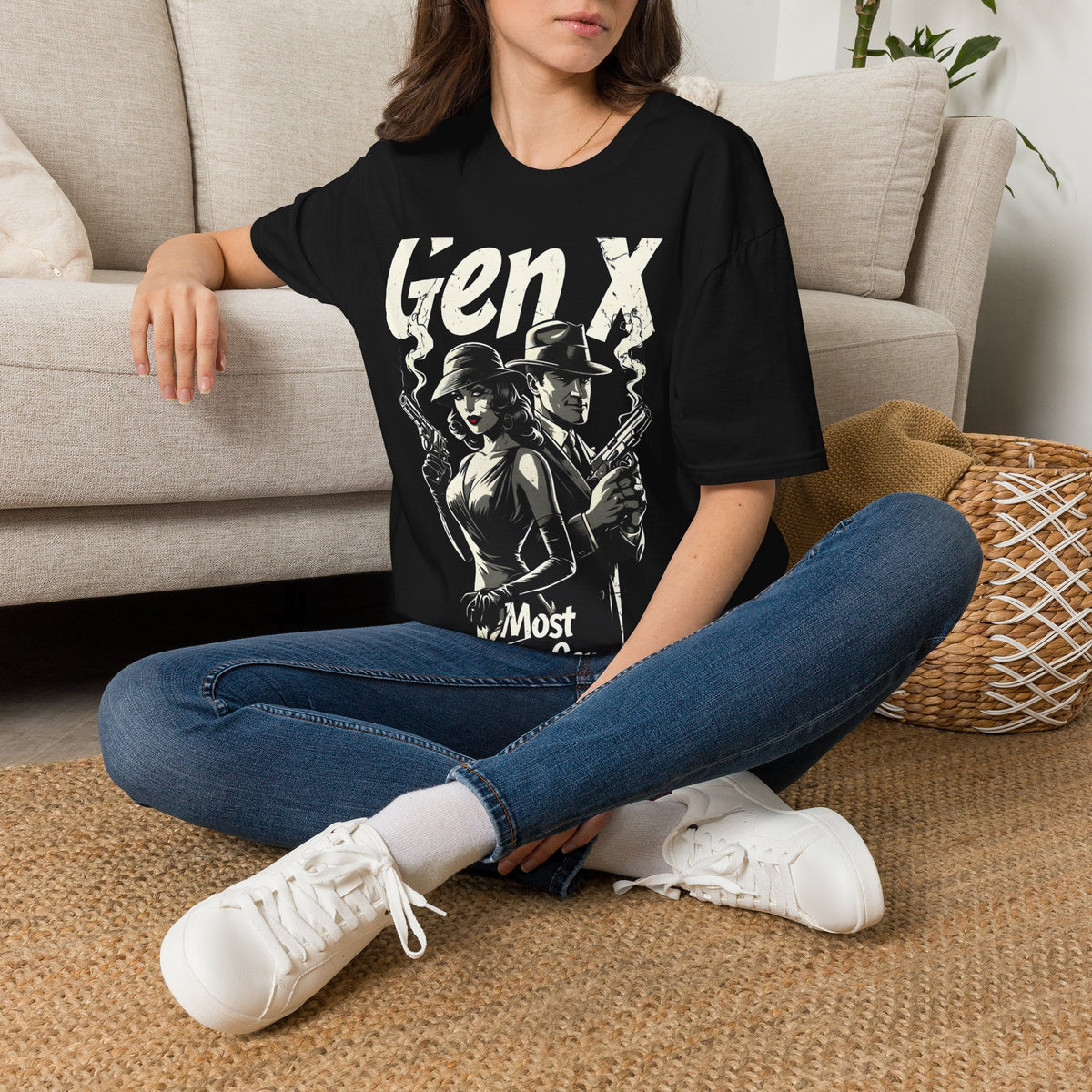 A woman stting on a rug on the floor next to a beige couch wearing a black t-shirt with a graphic of a man and woman standing back to back holding smoking guns with the text "GEN X MOST DANGEROUS GENERATION" wearing jeans and white sneakers.
