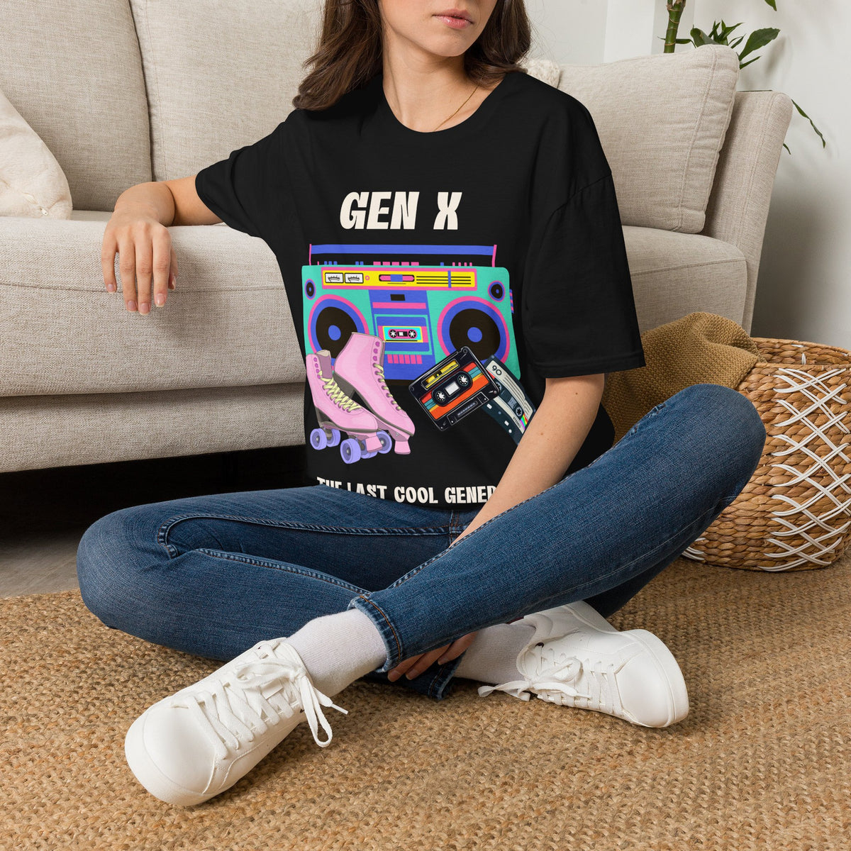 A woman sitting cross-legged on a woven rug in front of a beige sofa, wearing a black graphic T-shirt with the text 'GEN X THE COOLEST GENERATION' and a colorful retro boombox/roller skate design, blue jeans, and white sneakers.