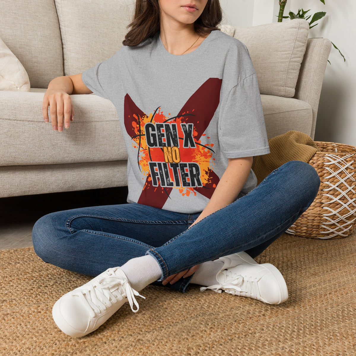 A woman sits cross-legged on a woven rug in front of a beige sofa, wearing a  gray graphic T-shirt with a large red 'X' design and the text 'GEN X NO FILTER' in black. She is also wearing blue jeans and white sneakers.