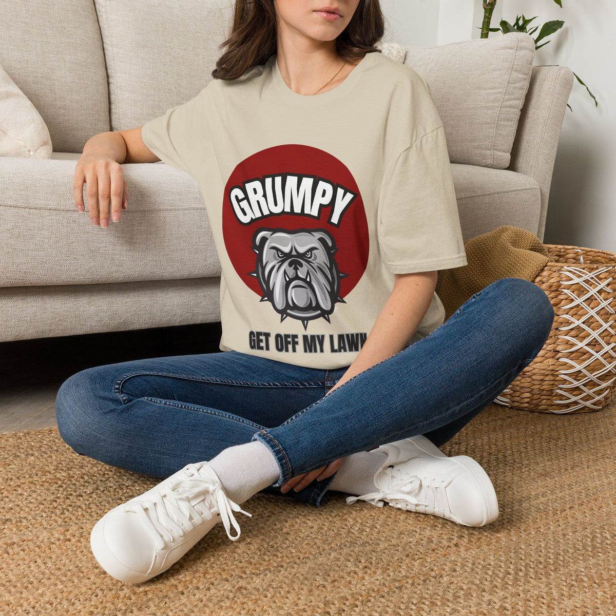 A woman sitting cross-legged on a woven rug in front of a light gray sofa, wearing a sand graphic t-shirt with a blue and yellow 'Member Completed' mug-themed badge, blue jeans, and white sneakers.