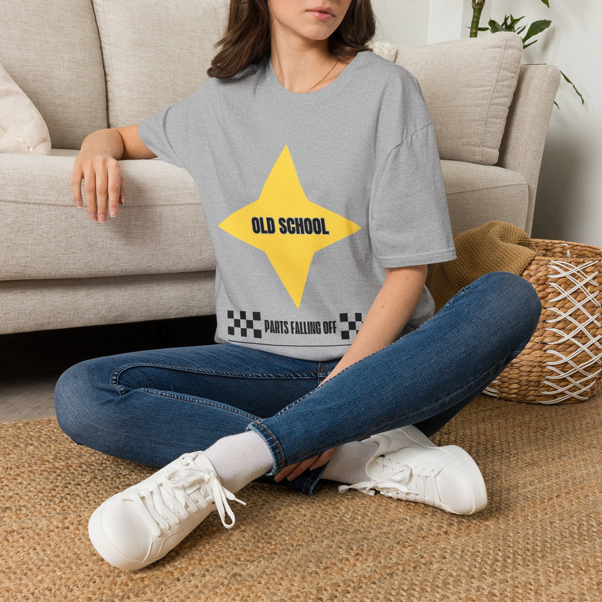 A woman sitting cross-legged on a woven rug in front of a beige sofa, wearing a gray graphic T-shirt with a large yellow star and the text 'OLD SCHOOL' and 'PARTS FALLING OFF'.