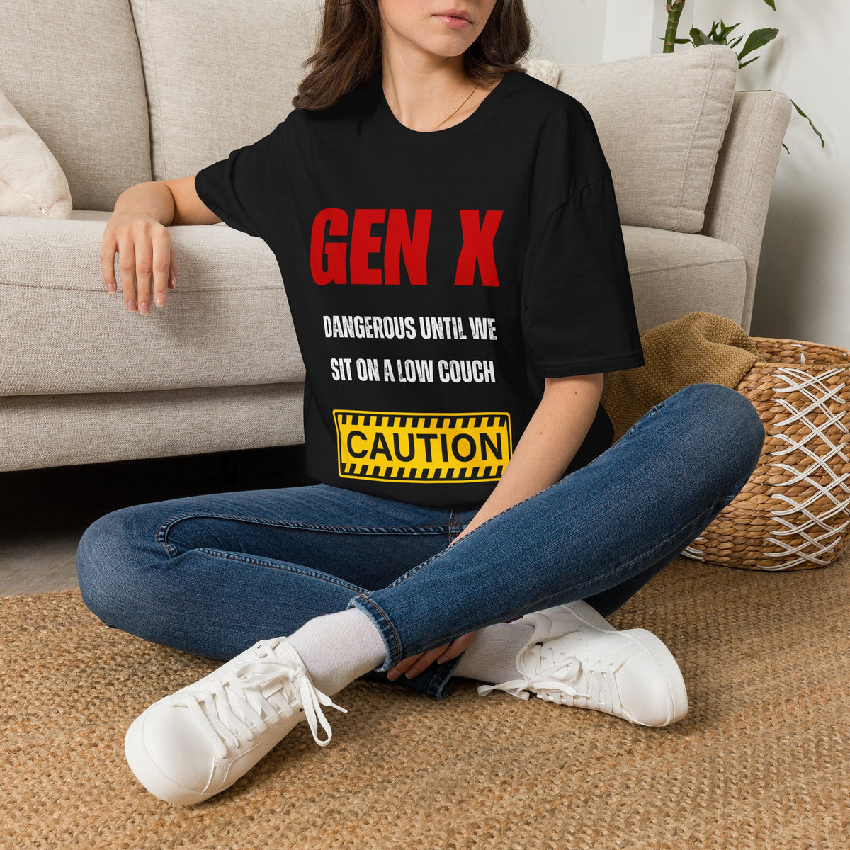 A woman sitting on a woven rug in front of a beige sofa, wearing a black graphic t-shirt with red and white text that reads 'GEN X DANGEROUS UNTIL WE SIT ON A LOW COUCH' and a yellow caution box graphic.