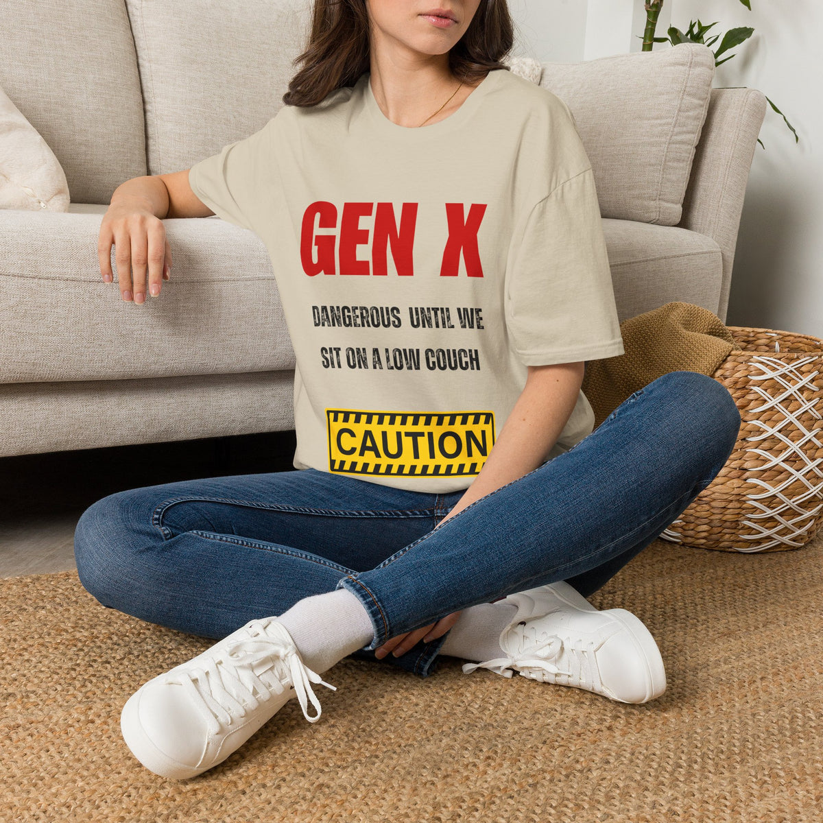 A woman sitting on a woven rug in front of a beige couch, wearing a sand graphic t-shirt with red and black text reading 'GEN X DANGEROUS UNTIL WE SIT ON A LOW COUCH' and a yellow caution-style box. She is also wearing blue jeans and white sneakers.