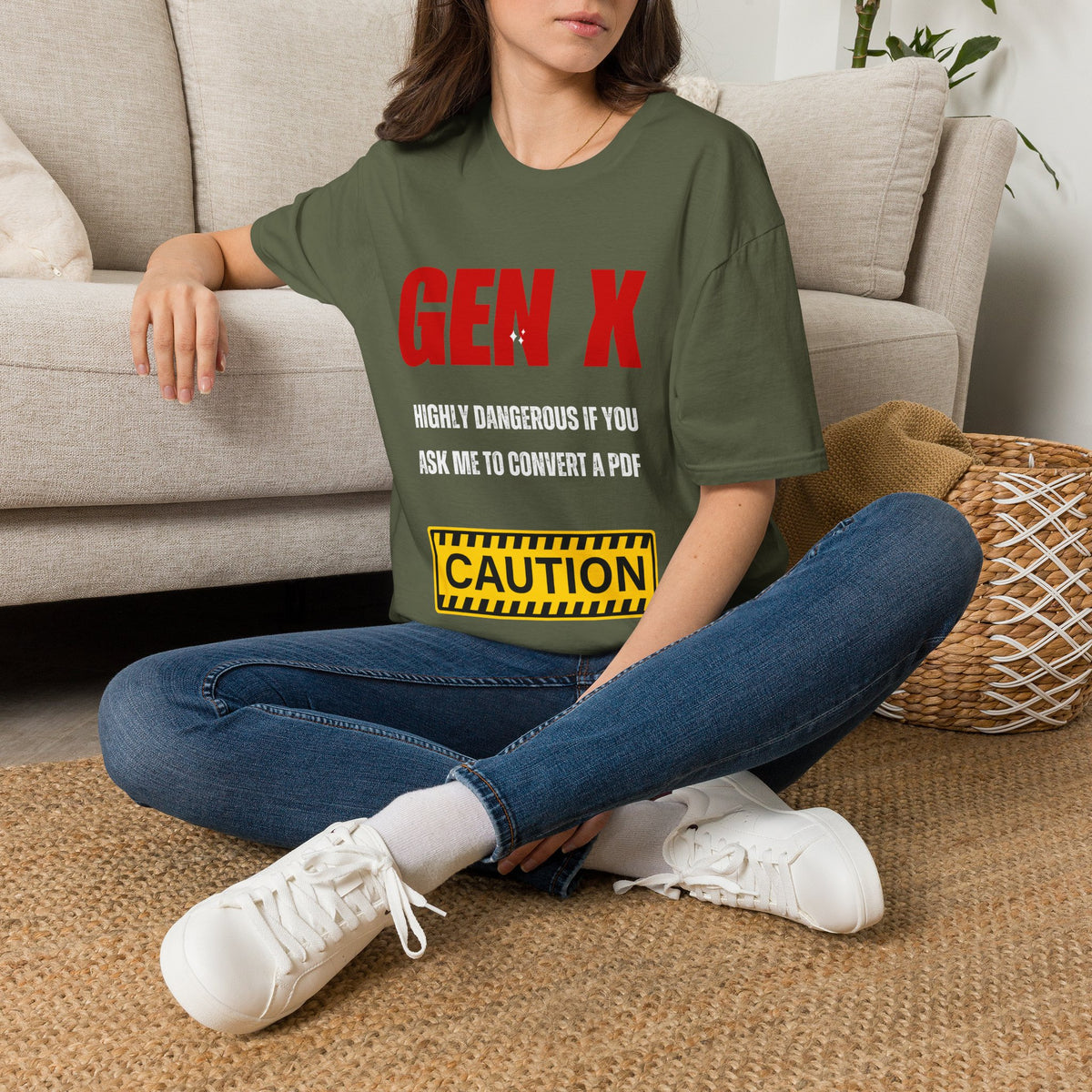 A woman sitting cross-legged on a woven rug in front of a beige sofa, wearing a green graphic T-shirt with red and white text reading 'GEN X' and a yellow caution-style box, paired with blue jeans and white sneakers.