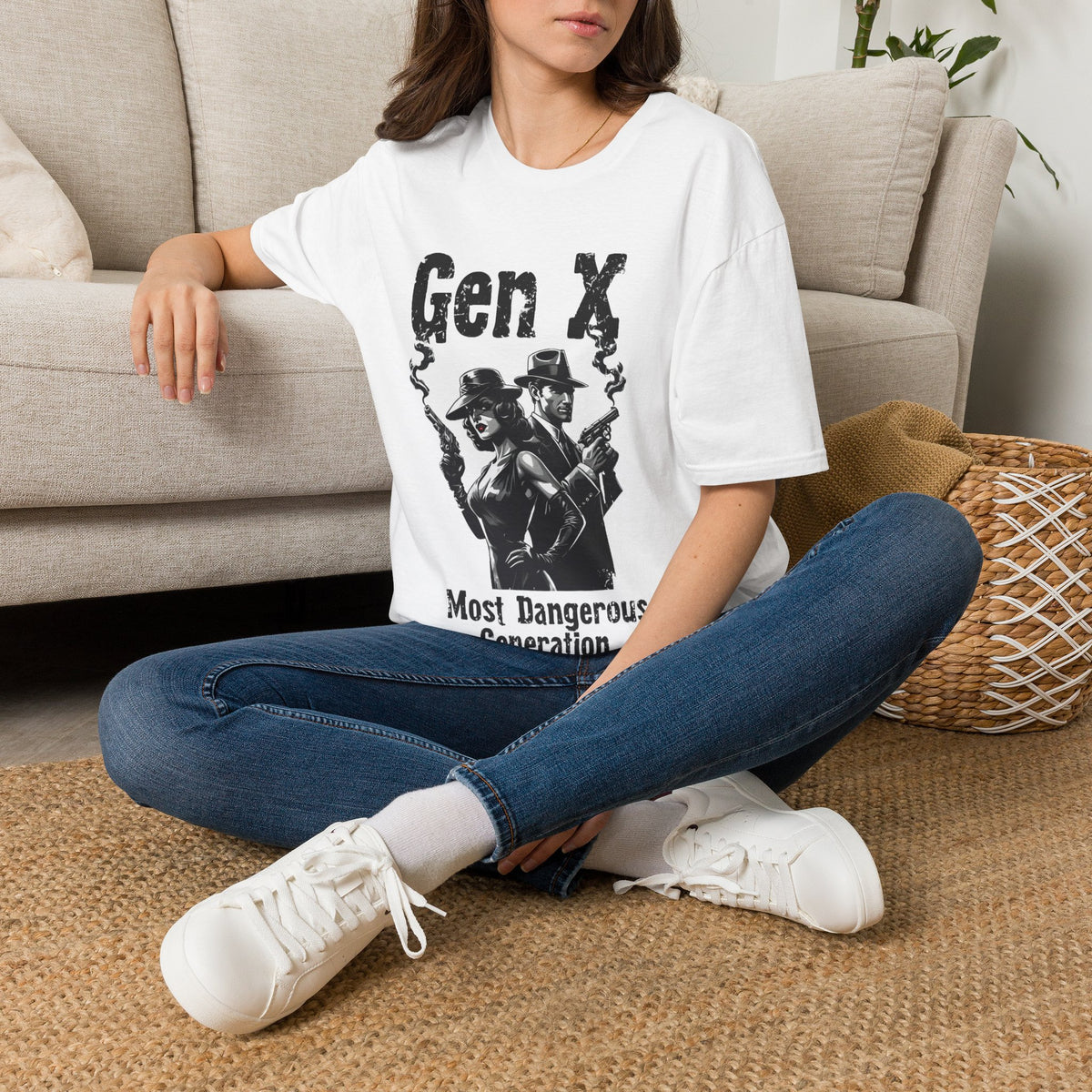 A woman sitting on a rug on the floor next to a biege couch, wearing a white t-shirt with the graphic of a woman and man standing back to back holding smoking guns and the text "GEN X MOIST DANGEROUS GENERATION".