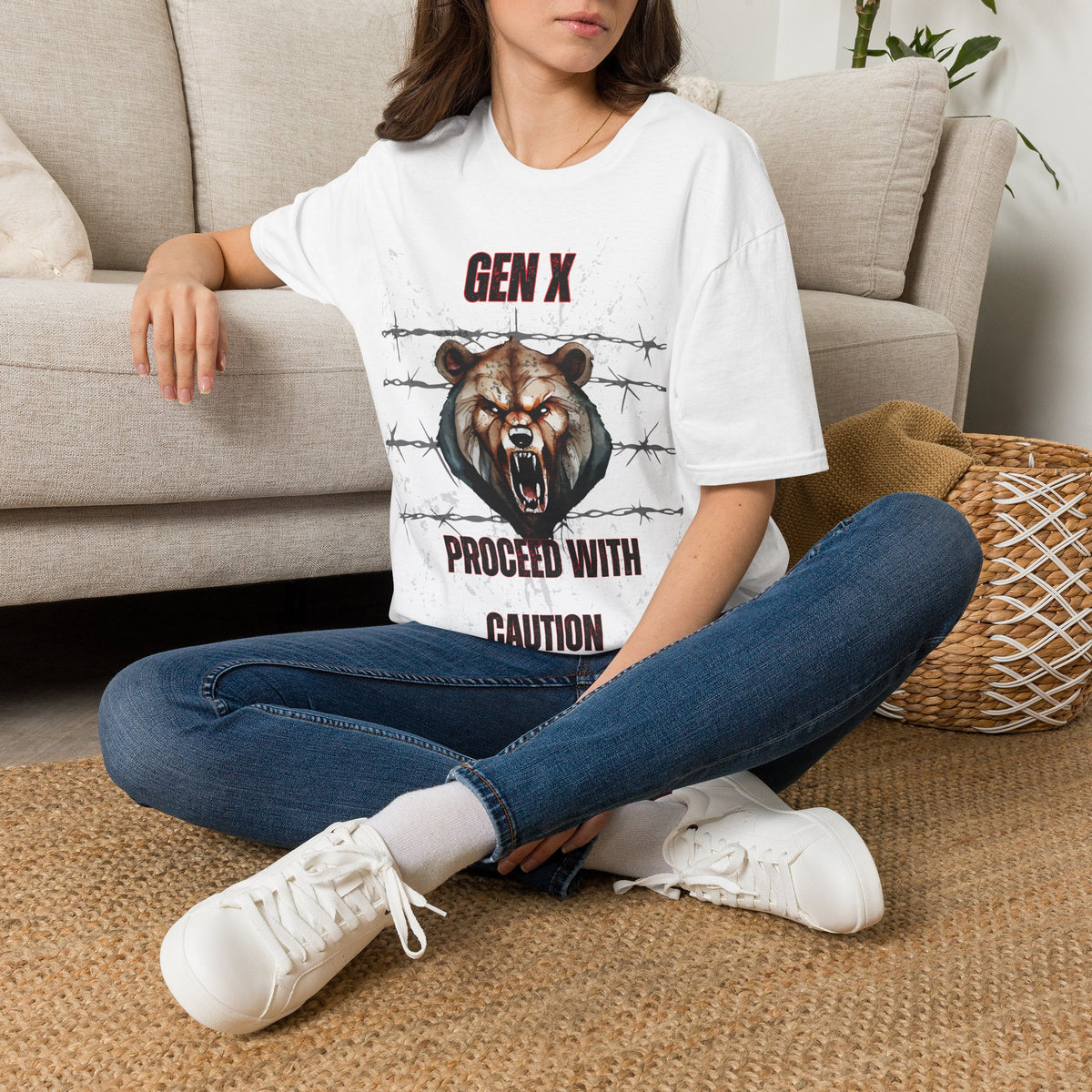 A woman sat on a rug net to a couch wearing a white graphic t-shirt featuring a roaring bear illustration and the text 'GEN X PROCEED WITH CAUTION'. 