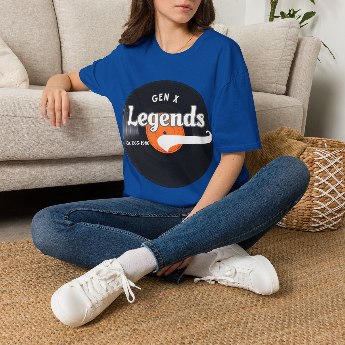 A woman sitting cross-legged on a woven rug in front of a beige sofa, wearing a royal blue graphic T-shirt with a vinyl record that reads 'GEN X Legends est 1965-1980' 