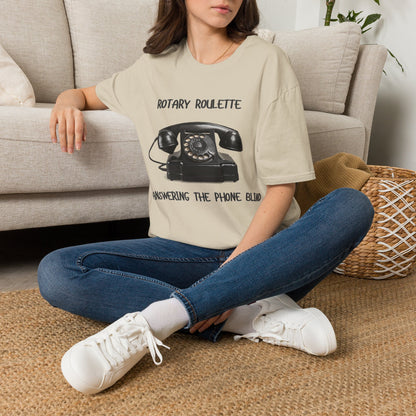 A woman sat on a rug next to a couch wearing a sand graphic t-shirt featuring a black rotary telephone and the text 'ROTARY ROULETTE' and 'ANSWERING THE PHONE BLIND'. 
