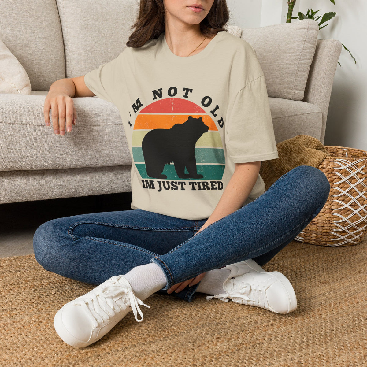 A woman sitting cross-legged on a woven rug in front of a light beige sofa, wearing an oversized sand graphic T-shirt with a bear silhouette and the text 'I'M NOT OLD I'M JUST TIRED', blue jeans, and white sneakers.