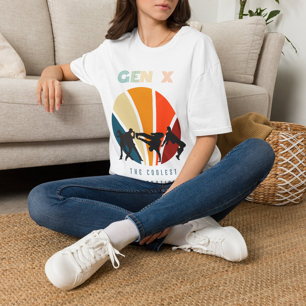 A woman sitting cross-legged on a woven rug in front of a beige sofa, wearing a white graphic T-shirt with a colorful retro sun design and the text 'GEN X' and 'THE COOLEST GENERATION'.