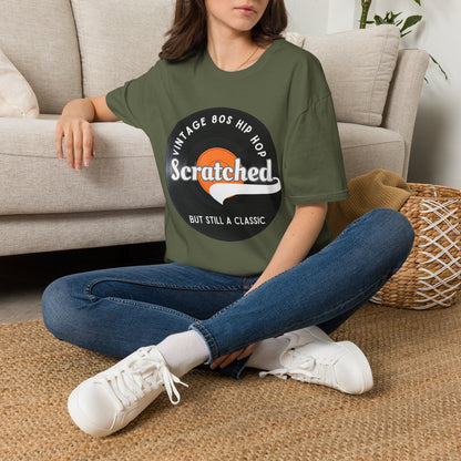 A woman sitting cross-legged on a woven rug in front of a beige sofa, wearing a green graphic t-shirt with a circular 'Vintage BOS Hip Hop' design featuring the word 'Scratched' in white. She is also wearing blue jeans and white sneakers.