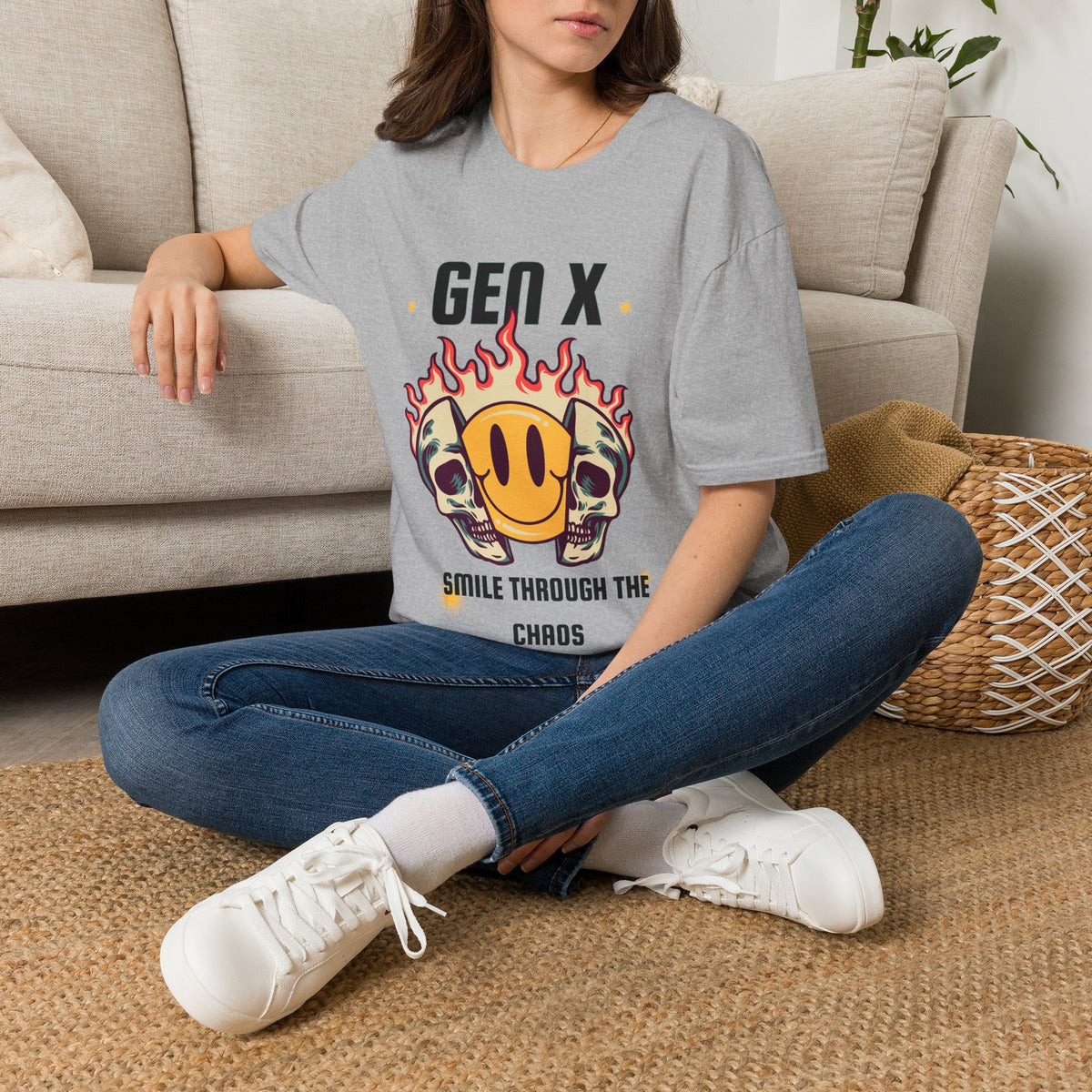 A woman sitting cross-legged on a woven rug in front of a beige sofa, wearing a light gray graphic T-shirt with a skull-and-smiley-face design and the text 'GEN X SMILE THROUGH THE CHAOS'.