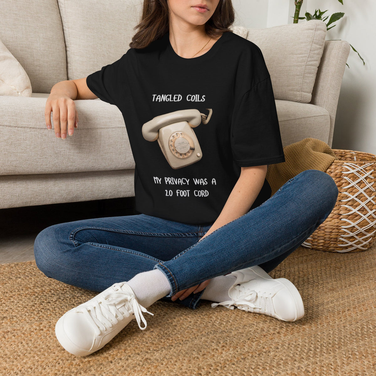 A woman sitting cross-legged on a woven rug in front of a beige sofa, wearing a black graphic T-shirt with white text and a vintage rotary phone illustration, blue jeans, and white sneakers.