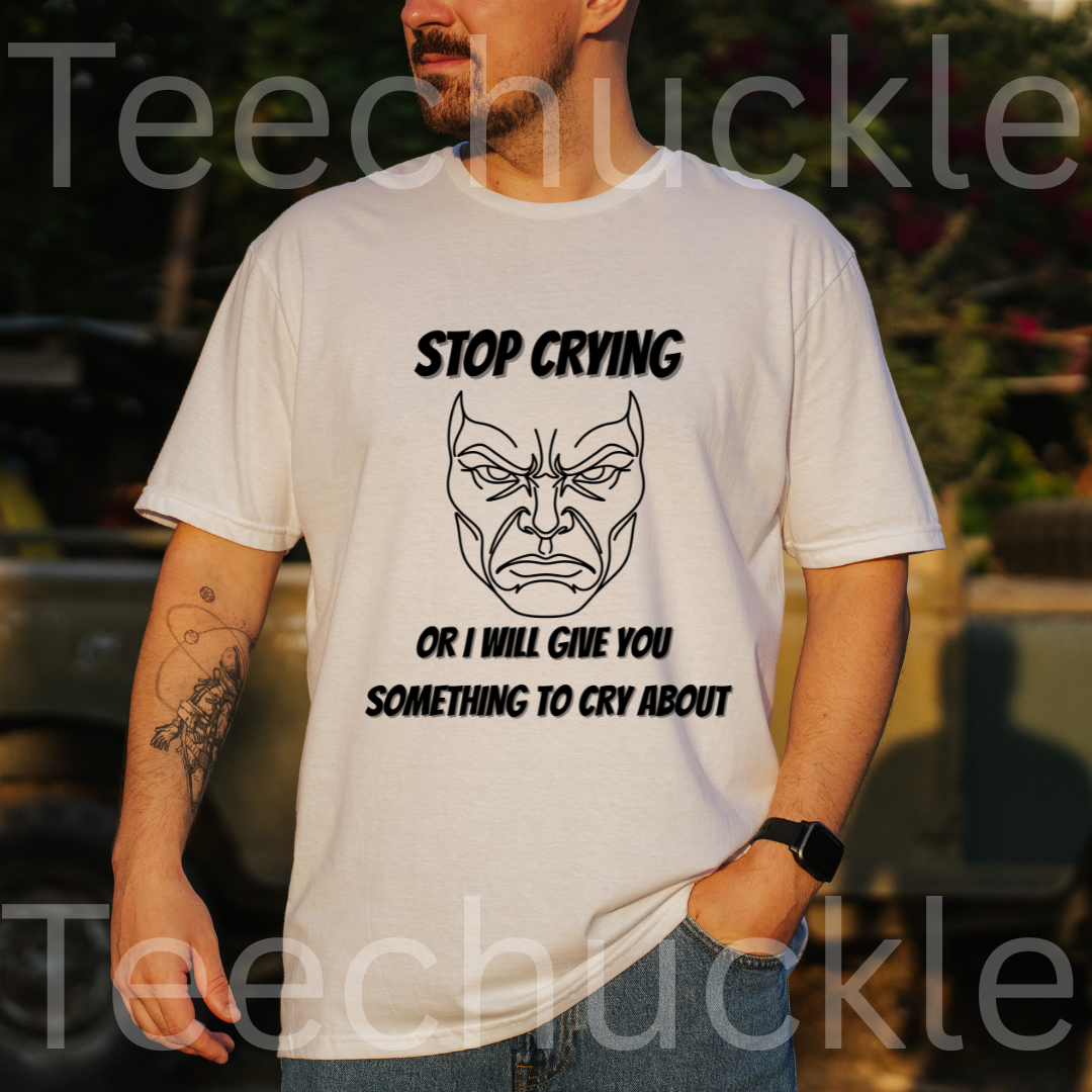 A man wearing a white graphic t-shirt with a black angry face and the text 'STOP CRYING OR I WILL GIVE YOU SOMETHING TO CRY ABOUT'. The background is a blurred outdoor setting with warm sunset lighting.