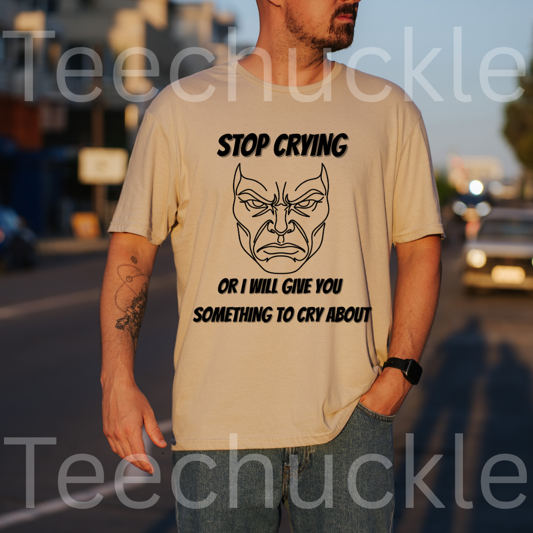 A man wearing a sand graphic t-shirt with a black printed angry face and the text 'STOP CRYING OR I WILL GIVE YOU SOMETHING TO CRY ABOUT', set against a blurred street background at sunset. 