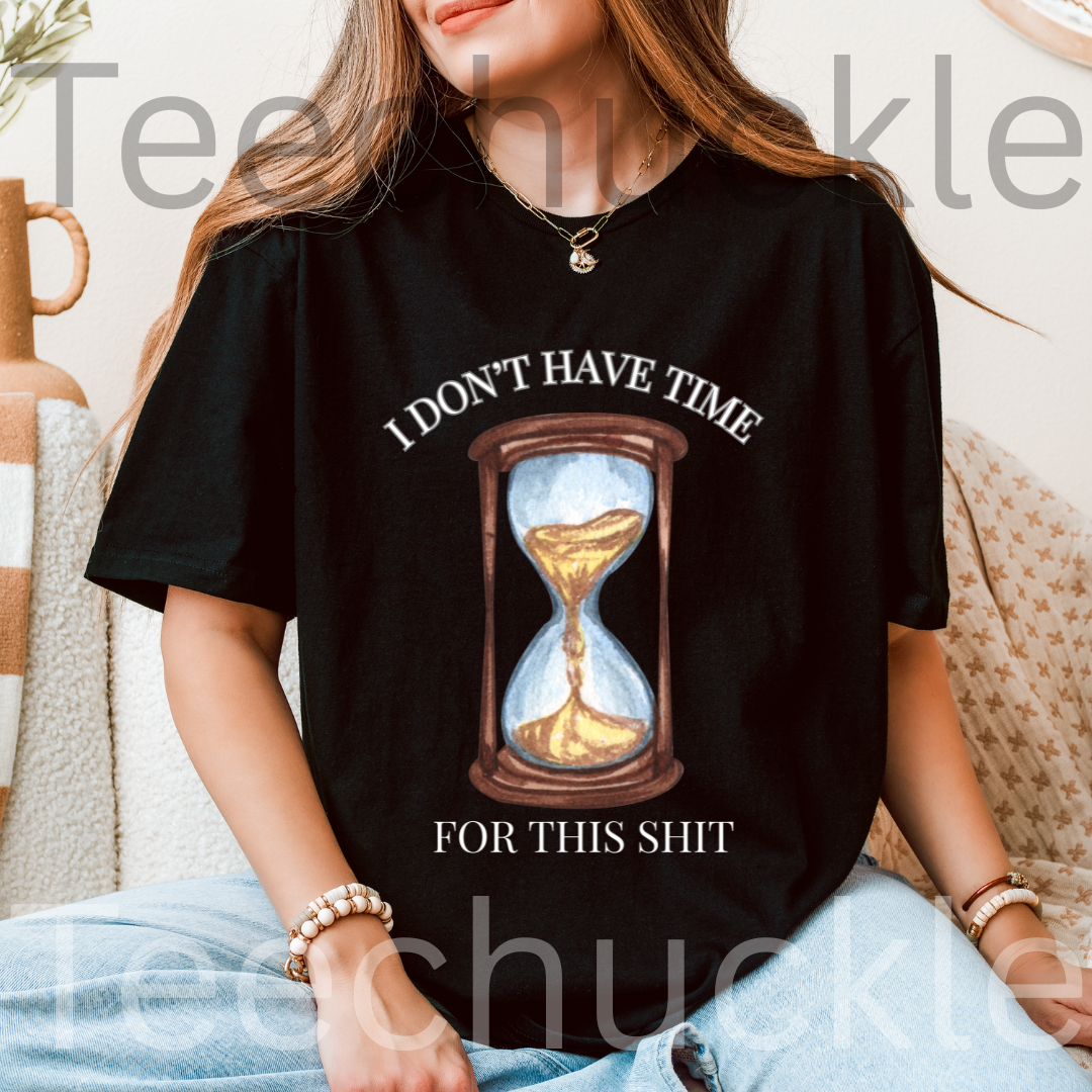 A woman wearing a black graphic t-shirt with an hourglass illustration,  and text " I DON'T HAVE TIME FOR THIS SHIT" paired with light blue jeans in a cozy indoor setting.