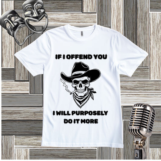 A white short-sleeve graphic T-shirt laid flat on a wood plank background, featuring a black skull wearing a cowboy hat and bandana with the text 'IF I OFFEND YOU I WILL PURPOSELY DO IT MORE.' Decorative vintage-style props, including a silver mask and microphone, appear around the shirt.