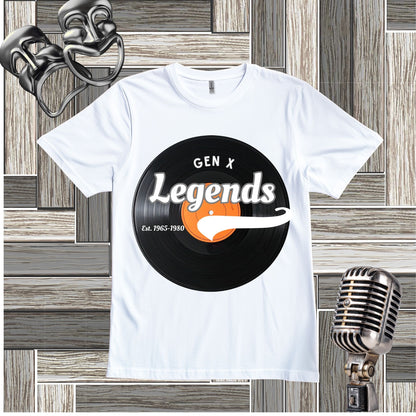 A white short-sleeve t-shirt with a retro graphic print featuring a black vinyl record, white text reading 'GEN X Legends', and an orange accent. The shirt is displayed on a gray wood-paneled background with vintage microphone and mask props.