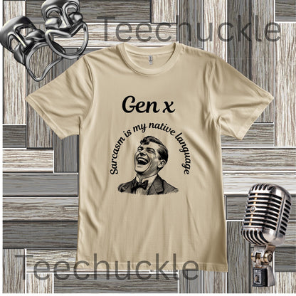 A sand graphic t-shirt featuring a black print of a laughing man and the text 'Gen x' and 'Sarcasm is my native language' arched around him. The t-shirt is displayed on a gray wood-plank background with vintage-style props including a microphone, a comedy masks.