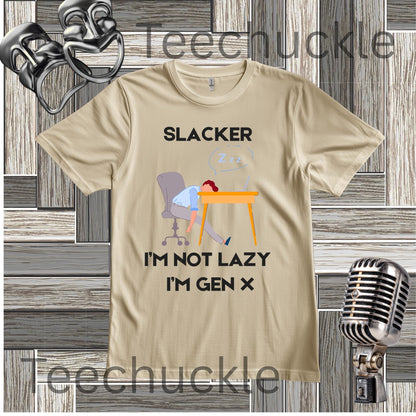 A sand graphic t-shirt featuring a cartoon illustration of a person sleeping at a desk with the text 'SLACKER I'M NOT LAZY I'M GEN X'. The t-shirt is displayed on a gray wood panel background with vintage-style microphones 