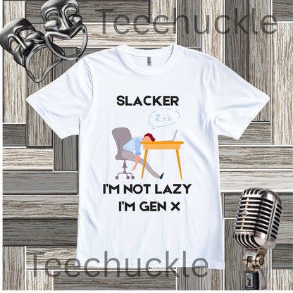 A white graphic t-shirt featuring a cartoon illustration of a person sleeping at a desk with the text 'SLACKER I'M NOT LAZY I'M GEN X'. The t-shirt is displayed on a gray wood-plank background with a vintage microphone, 