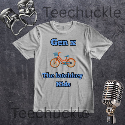 A gray graphic t-shirt featuring the text 'Gen x' and 'The latchkey Kids' in blue, with an orange bicycle in the center. The t-shirt is displayed on a dark textured background with vintage silver props (comedy masks and a microphone)